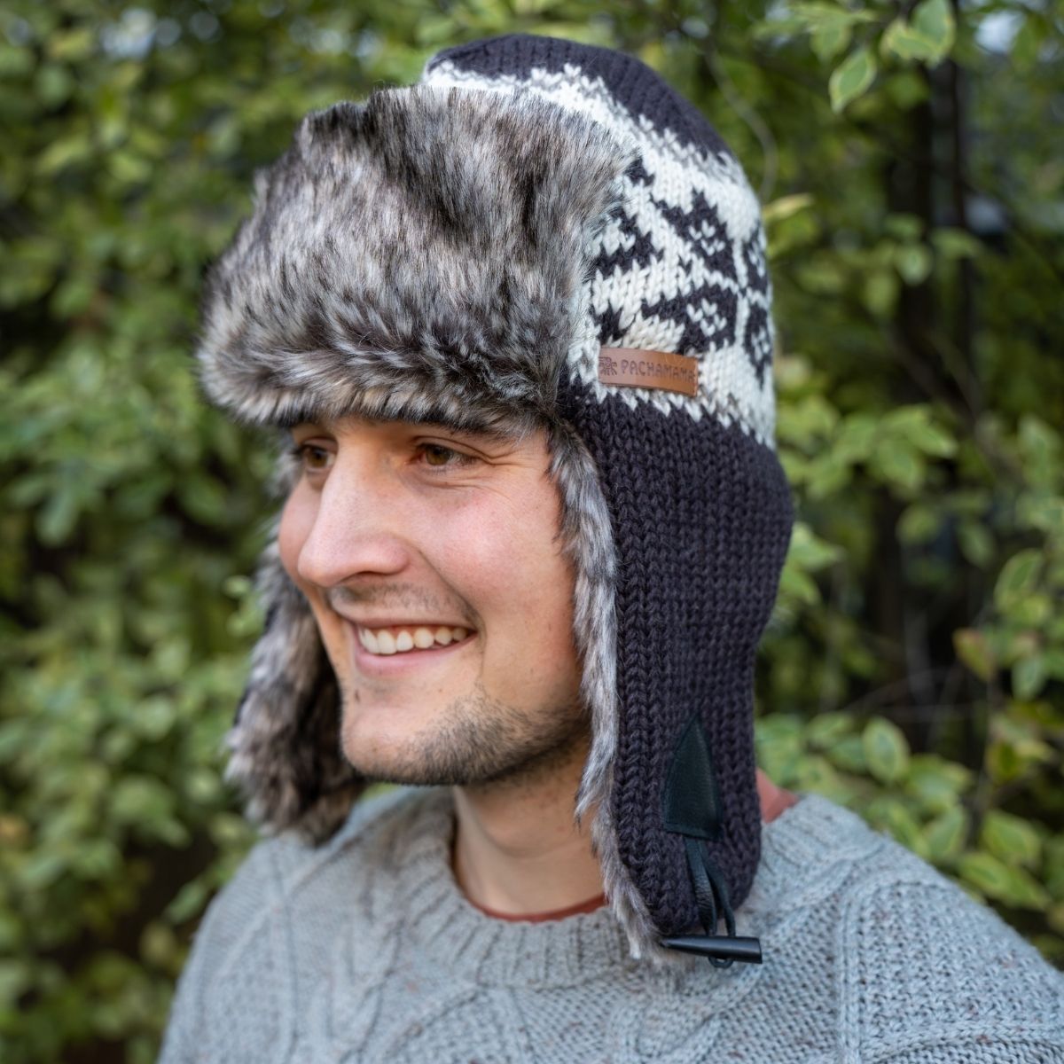 A man wearing a dark navy and white knit winter trapper hat with faux fur ear flaps, photographed outdoors against a blurred green foliage background.