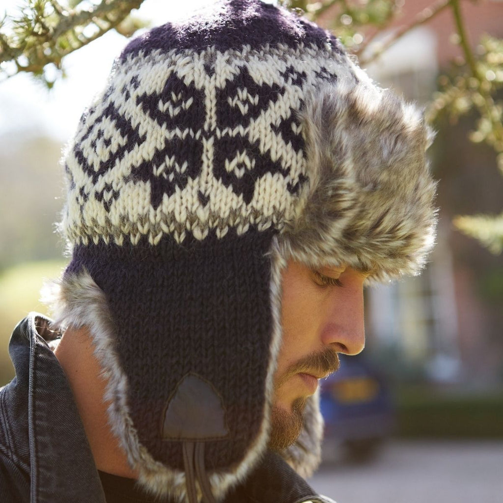A man wearing a dark navy knit trapper hat with a cream and navy Fair Isle pattern on the crown, a faux fur trim, and ear flaps, photographed outdoors in bright natural light.
