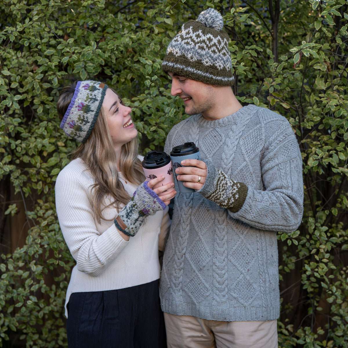 A couple outdoors in front of green foliage, wearing matching knit winter accessories including a patterned beanie, matching knit gloves, and a cream knit sweater. The man wears a gray cable-knit sweater and beige pants, while the woman wears a cream knit sweater and dark pants.