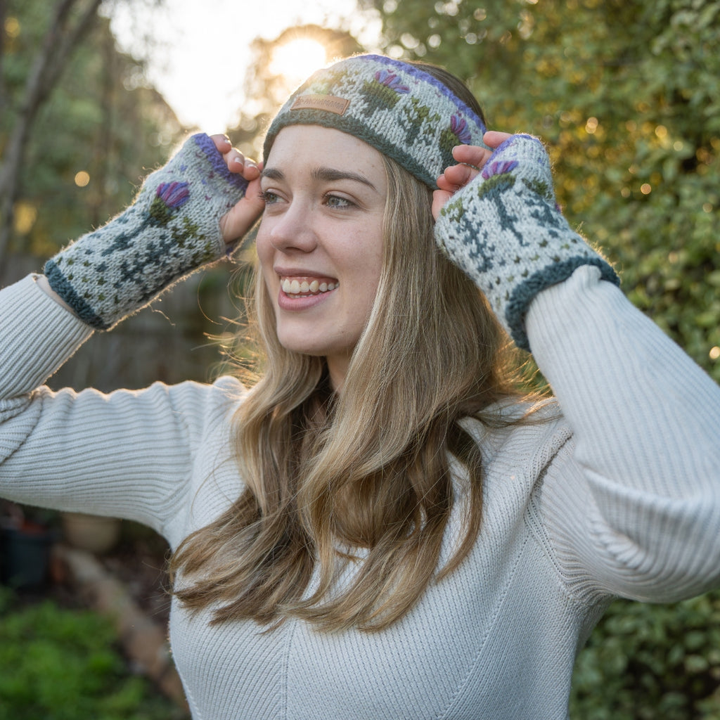 A woman outdoors wearing a knitted beanie and matching fingerless gloves, with her hands adjusting the beanie. The knitwear has a light gray base with purple, green, and blue floral and geometric patterns.