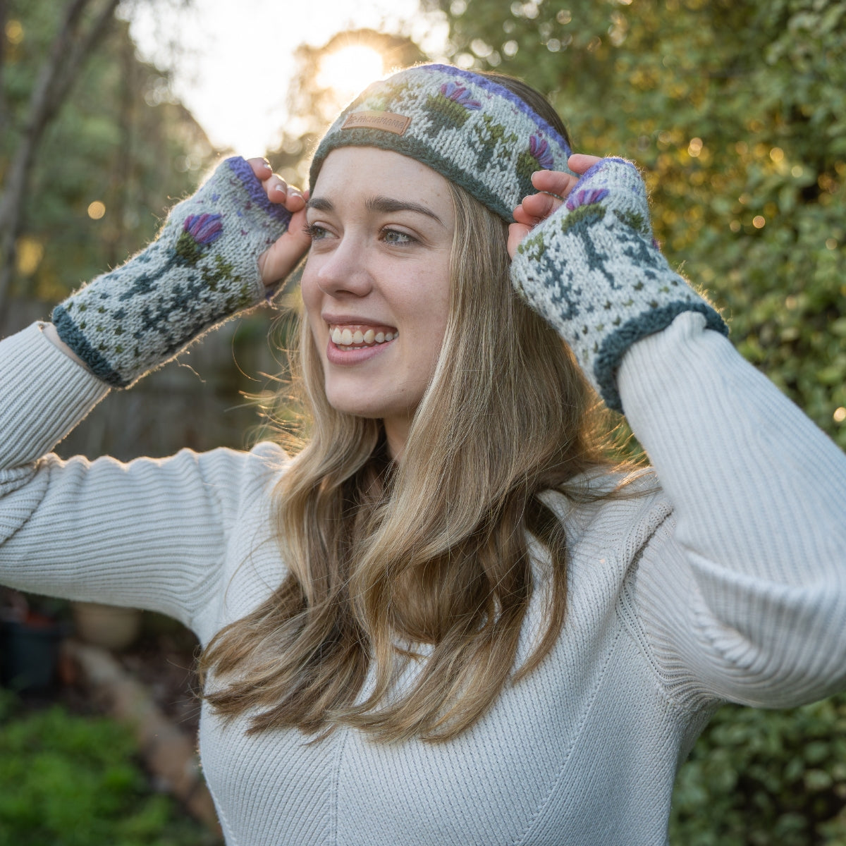A woman outdoors wearing a knitted beanie and matching fingerless gloves, with her hands adjusting the beanie. The knitwear has a light gray base with purple, green, and blue floral and geometric patterns.