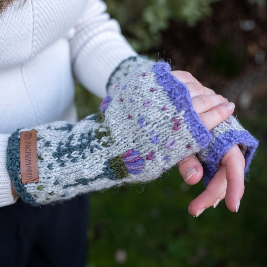A person outdoors wearing a pair of knit fingerless gloves. The gloves are light gray with purple ribbed cuffs, purple and pink floral accents, and a brown leather brand tab reading 'Pikimurphy'.