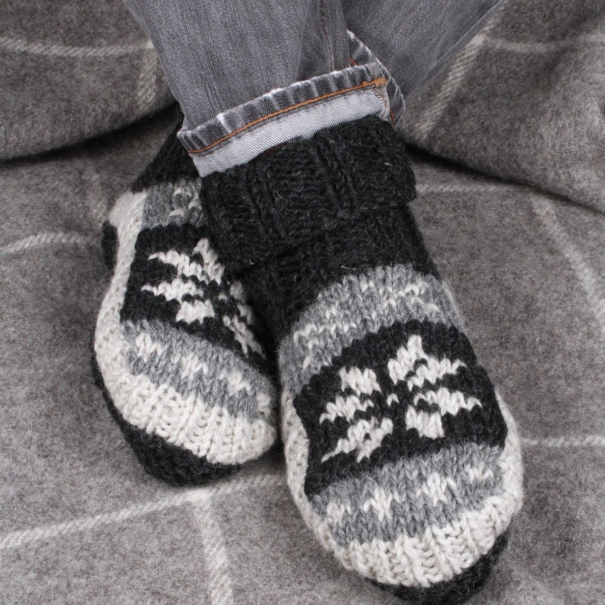 A close-up of a person's feet wearing black, gray, and white knit slipper socks with a snowflake pattern, paired with gray jeans, sitting on a gray plaid blanket.