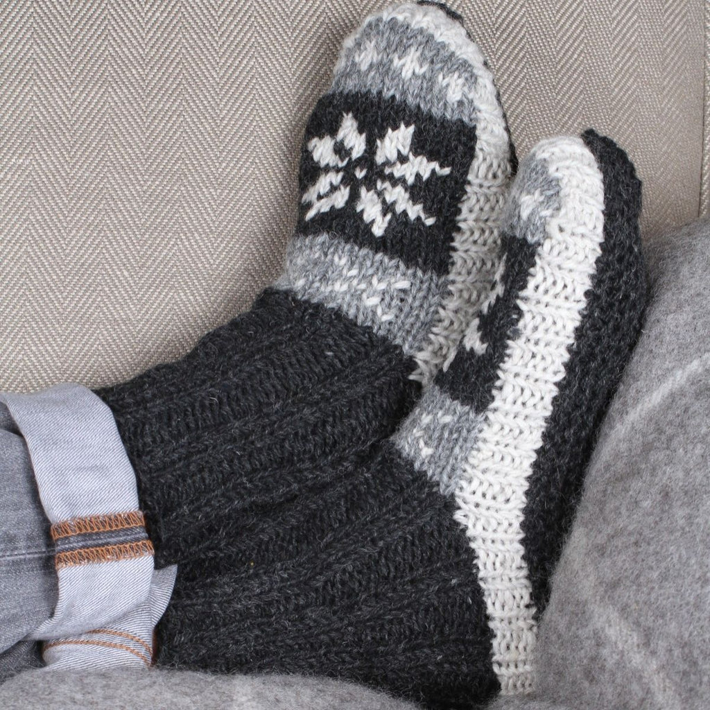 A close-up of a person's feet wearing chunky knit winter socks with a dark gray and white Nordic snowflake pattern, resting on a gray couch.