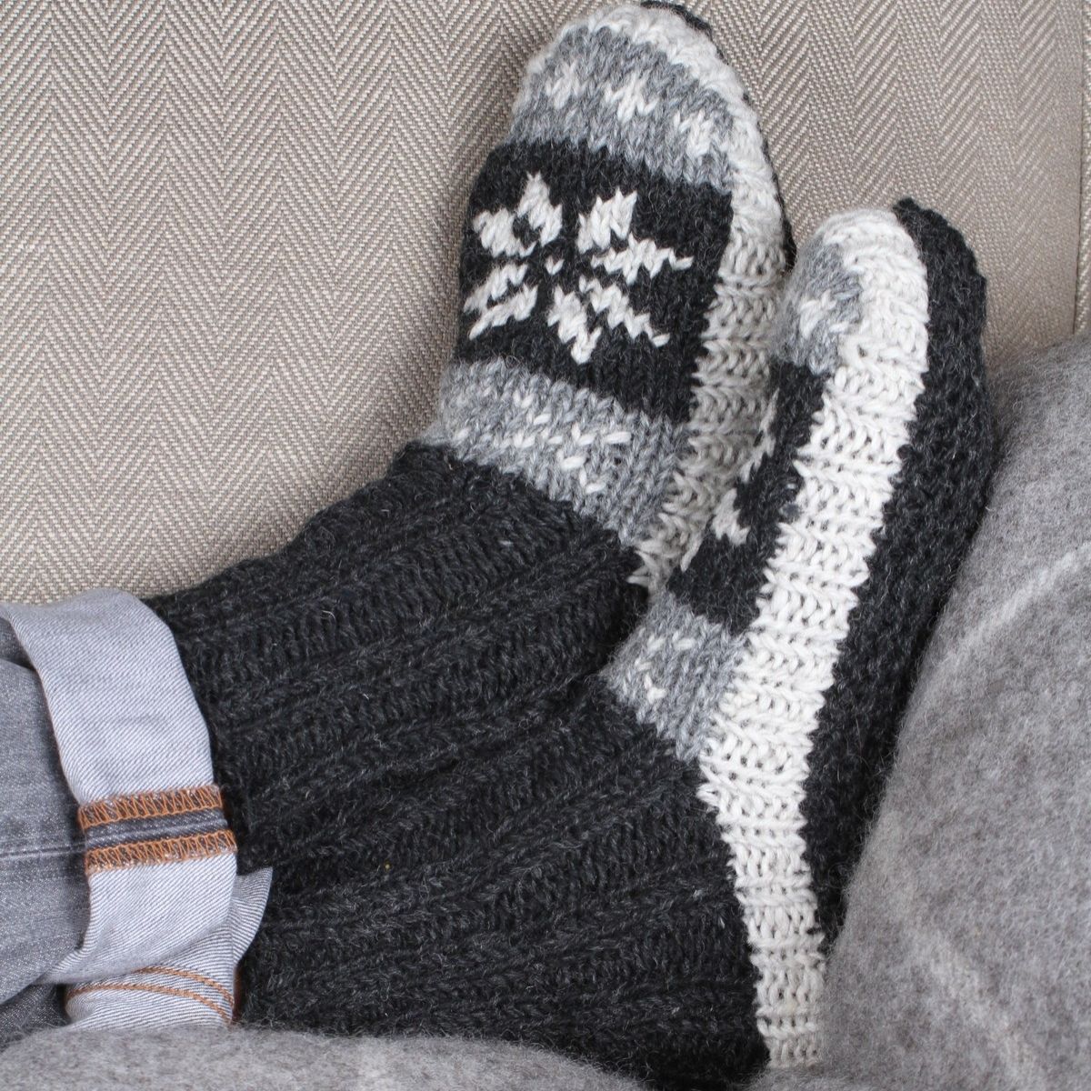 A close-up of a person's feet wearing chunky knit winter socks with a dark gray and white Nordic snowflake pattern, resting on a gray couch.