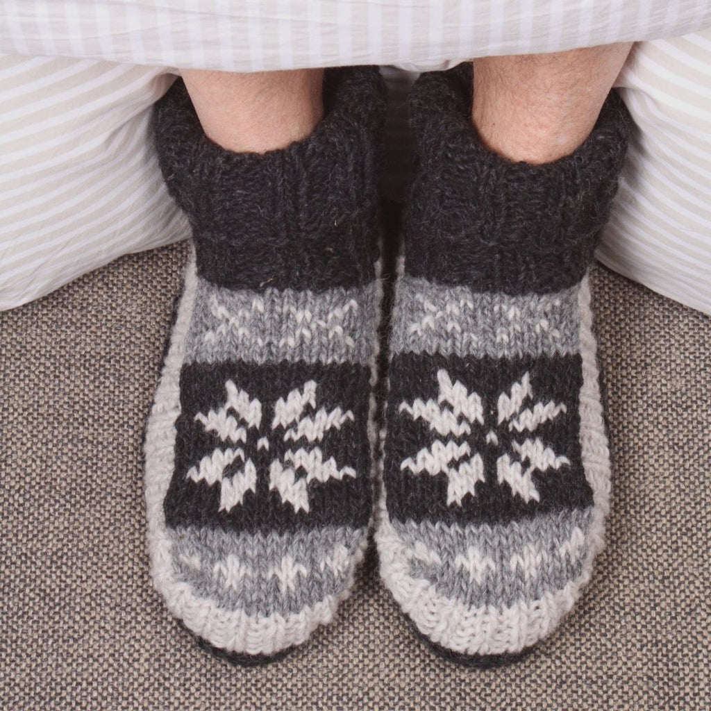 A close-up of a person's feet wearing cozy hand-knit slipper socks with a dark gray ribbed cuff, a gray and white snowflake pattern, and a white striped hem, resting on a textured gray carpet.