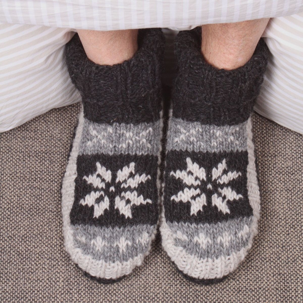 A close-up of a person's feet wearing cozy hand-knit slipper socks with a dark gray ribbed cuff, a gray and white snowflake pattern, and a white striped hem, resting on a textured gray carpet.