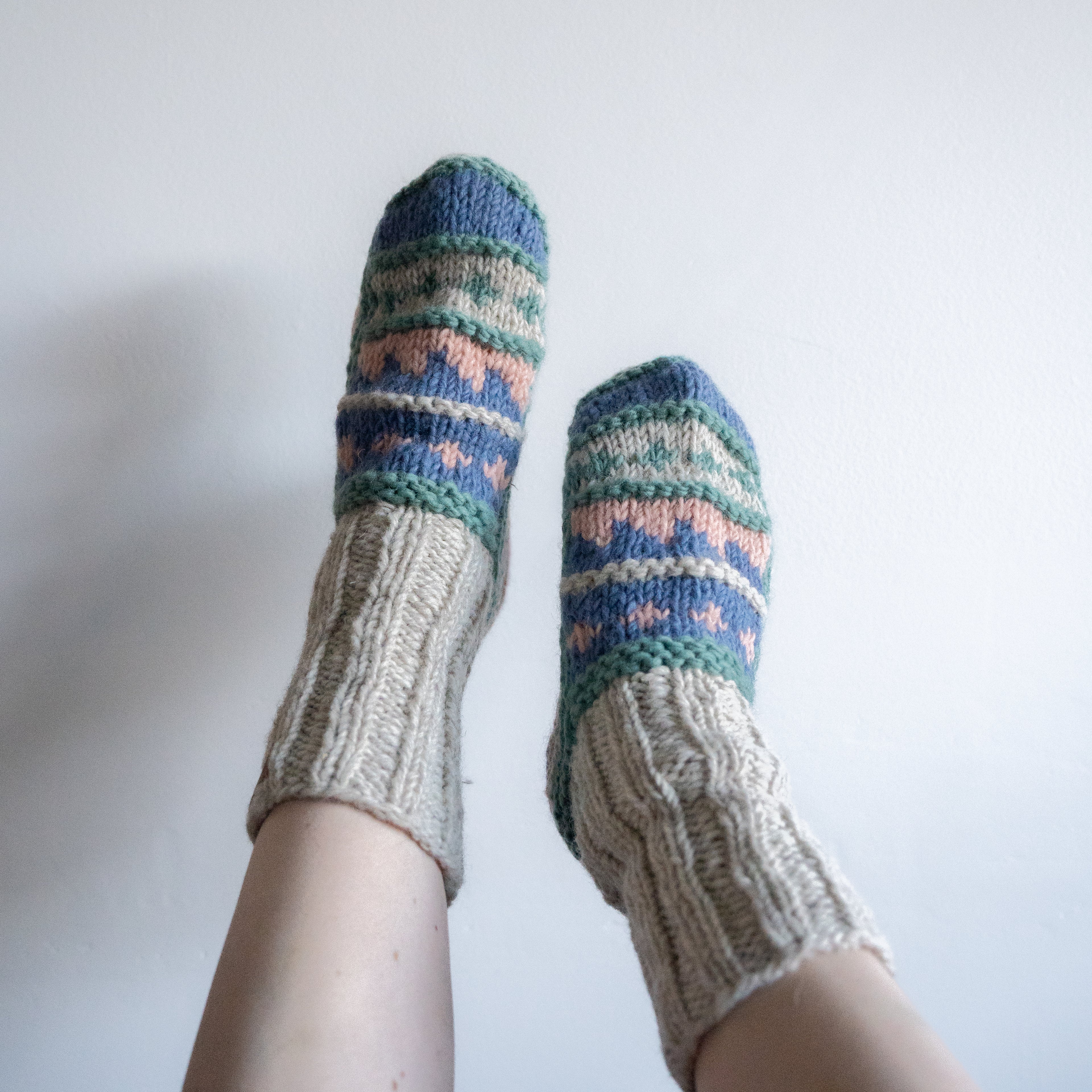A pair of hand-knit socks with a cream cable-knit leg section and a multicolor patterned foot section, shown being worn on feet against a plain white wall.
