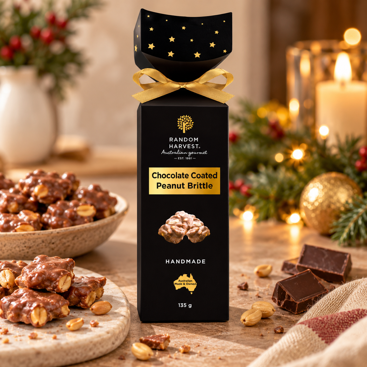A box of Random Harvest Chocolate Coated Peanut Brittle stands on a wooden table in a warm, festive setting with lit candles, pine branches, and extra brittle pieces nearby.