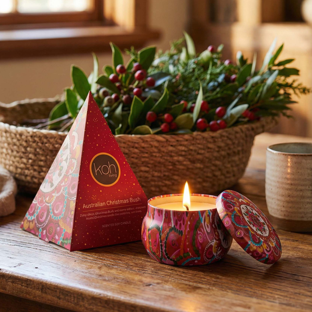 A lit candle in a colorful patterned tin sits on a wooden table beside its red pyramid-shaped box, with a woven basket of greenery in the background.