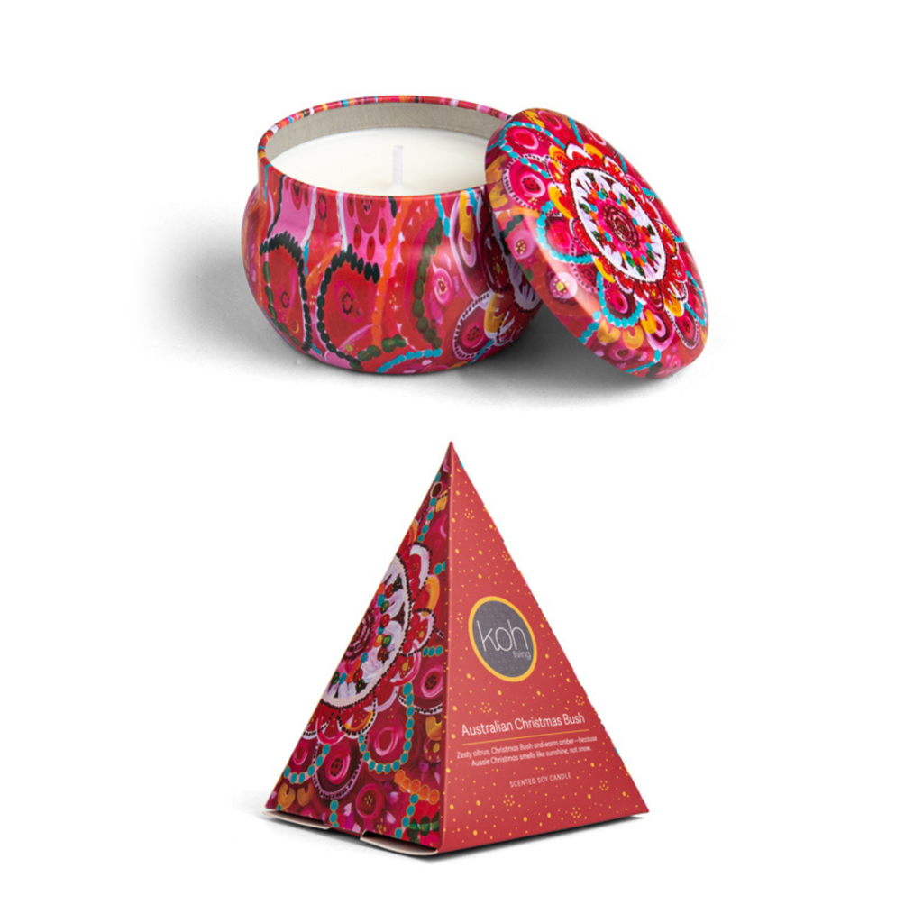 A colorful patterned tin candle shown open with the lid leaning against it, alongside its pyramid-shaped retail box. The candle and packaging feature a vivid red, pink, orange, and blue mandala-style design.