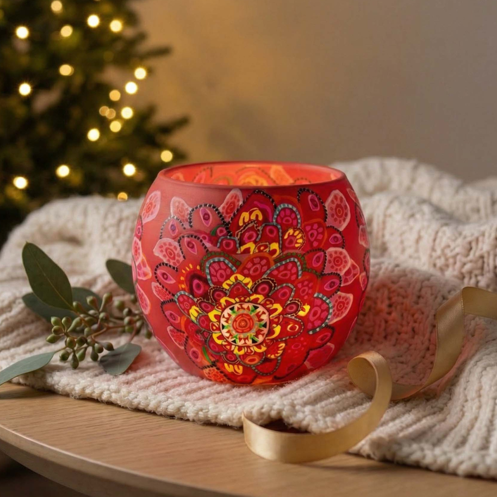 A red glass candle holder with a colorful mandala pattern, lit from within, styled on a cream knit blanket with a gold ribbon and eucalyptus sprig, with a blurred Christmas tree in the background.