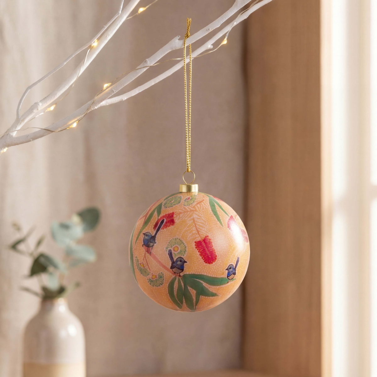A round decorative Christmas ornament with a warm peach background, hand-painted green leaves, red accents, and small blue birds, hanging from a gold string on a white branch with warm fairy lights.