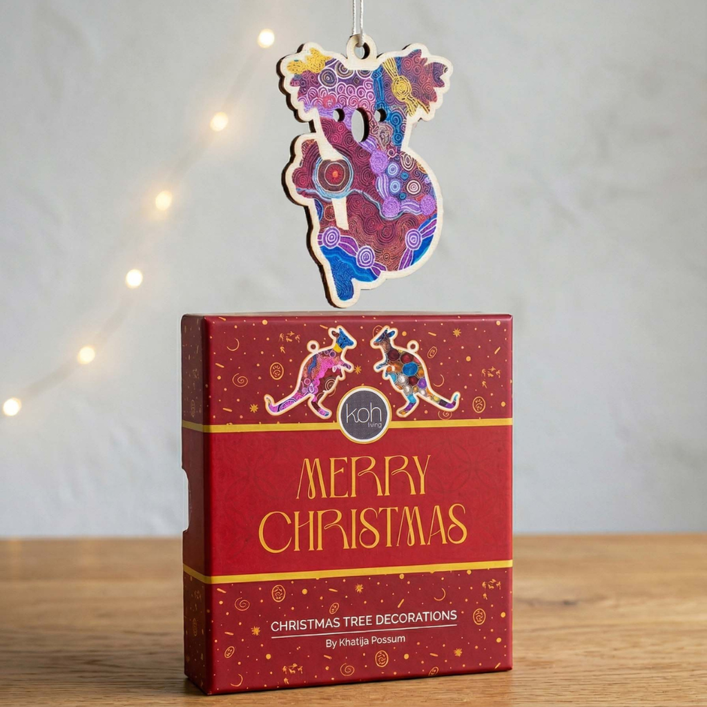 A colorful wooden Christmas ornament with intricate Aboriginal-inspired patterns hangs above a red gift box labeled 'MERRY CHRISTMAS' on a wooden surface against a softly blurred background with warm string lights.