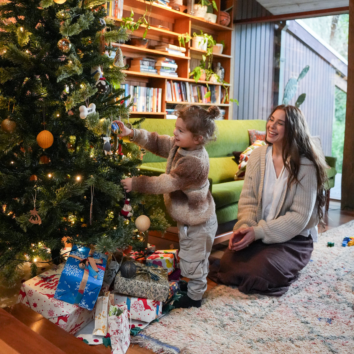 A young child in a brown fuzzy sweater and light pants decorates a lit Christmas tree while a woman kneels beside them in a cozy living room with bookshelves, a green sofa, and wrapped gifts under the tree.