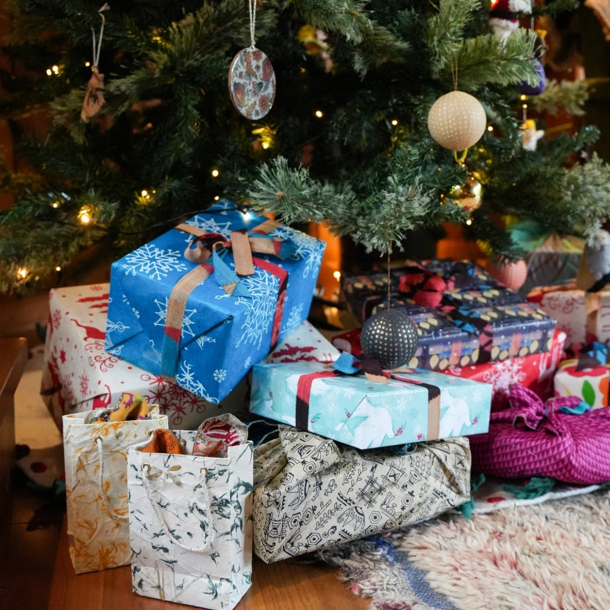 A collection of wrapped Christmas gifts and gift bags arranged under a decorated Christmas tree. The wrapping paper features various festive patterns including snowflakes, holiday trees, stripes, and textured designs in blue, white, red, green, and purple, accented with ribbons and bows.