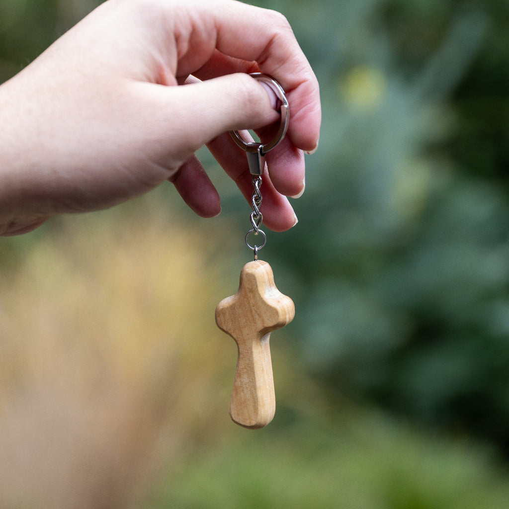 A hand holding a small wooden cross keychain with a silver metal split ring and chain against a blurred green outdoor background.