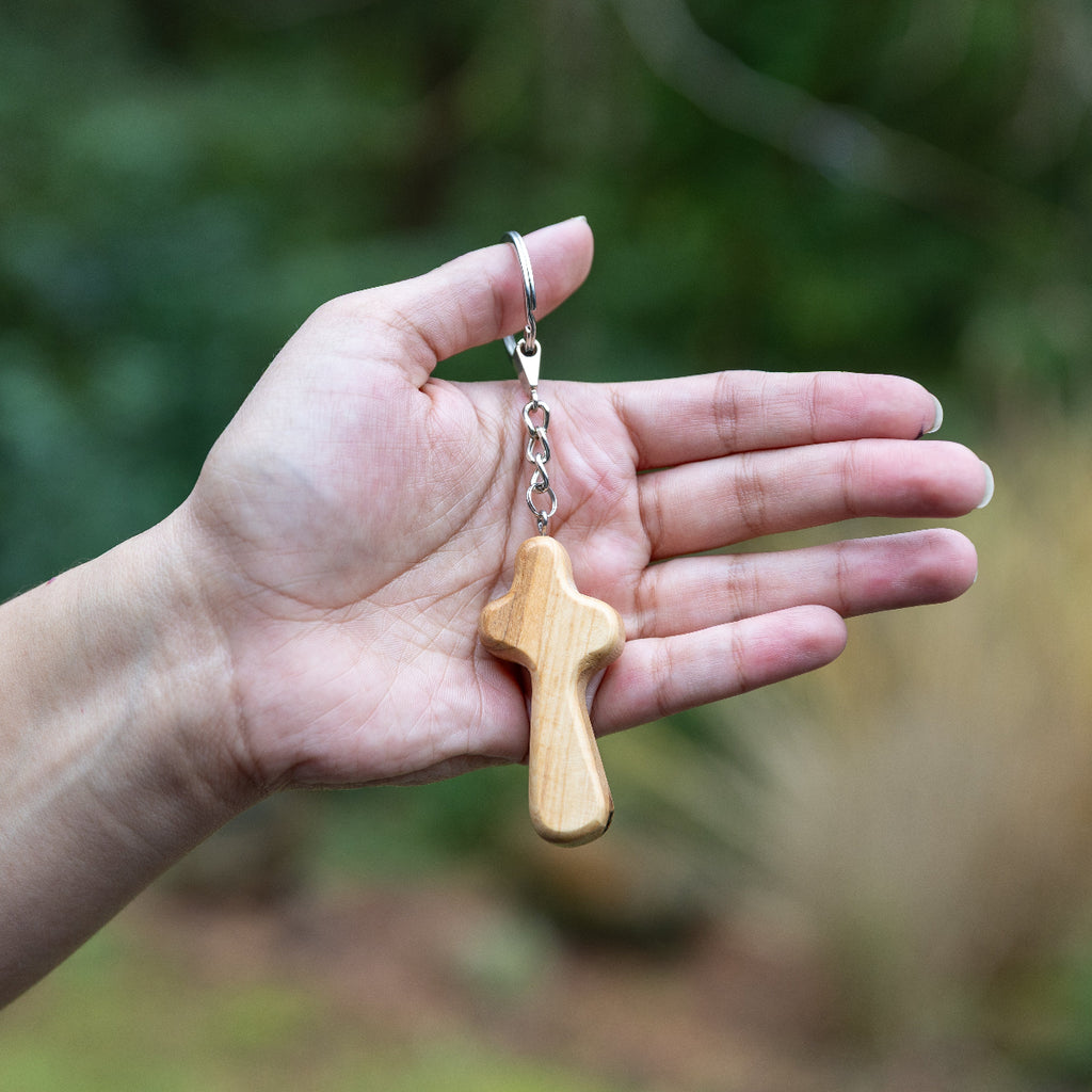 A small wooden cross keychain with a silver metal chain and split ring, held in a person's hand against a blurred outdoor green background.