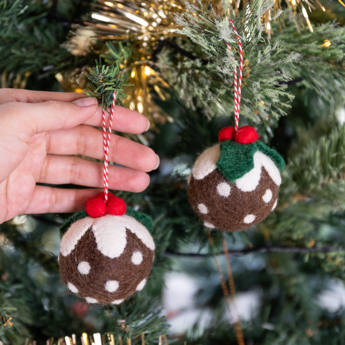Hand holding a brown felt Christmas pudding ornament with white polka dots, green felt leaves, and red berries, next to another hanging ornament on a Christmas tree.