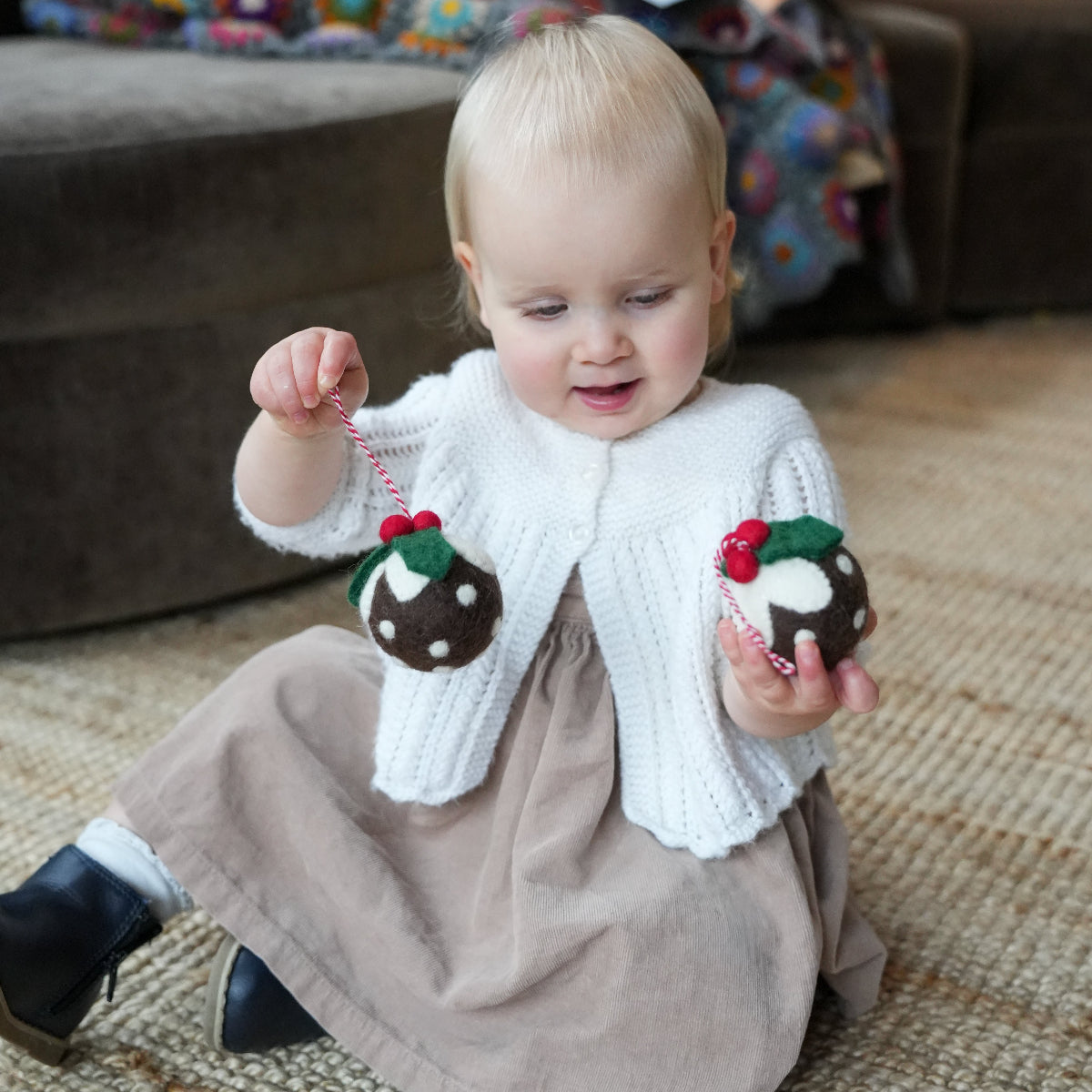 A toddler sitting on a woven rug indoors, wearing a white knit cardigan and taupe dress, while holding two small Christmas pudding ornaments with red berries.