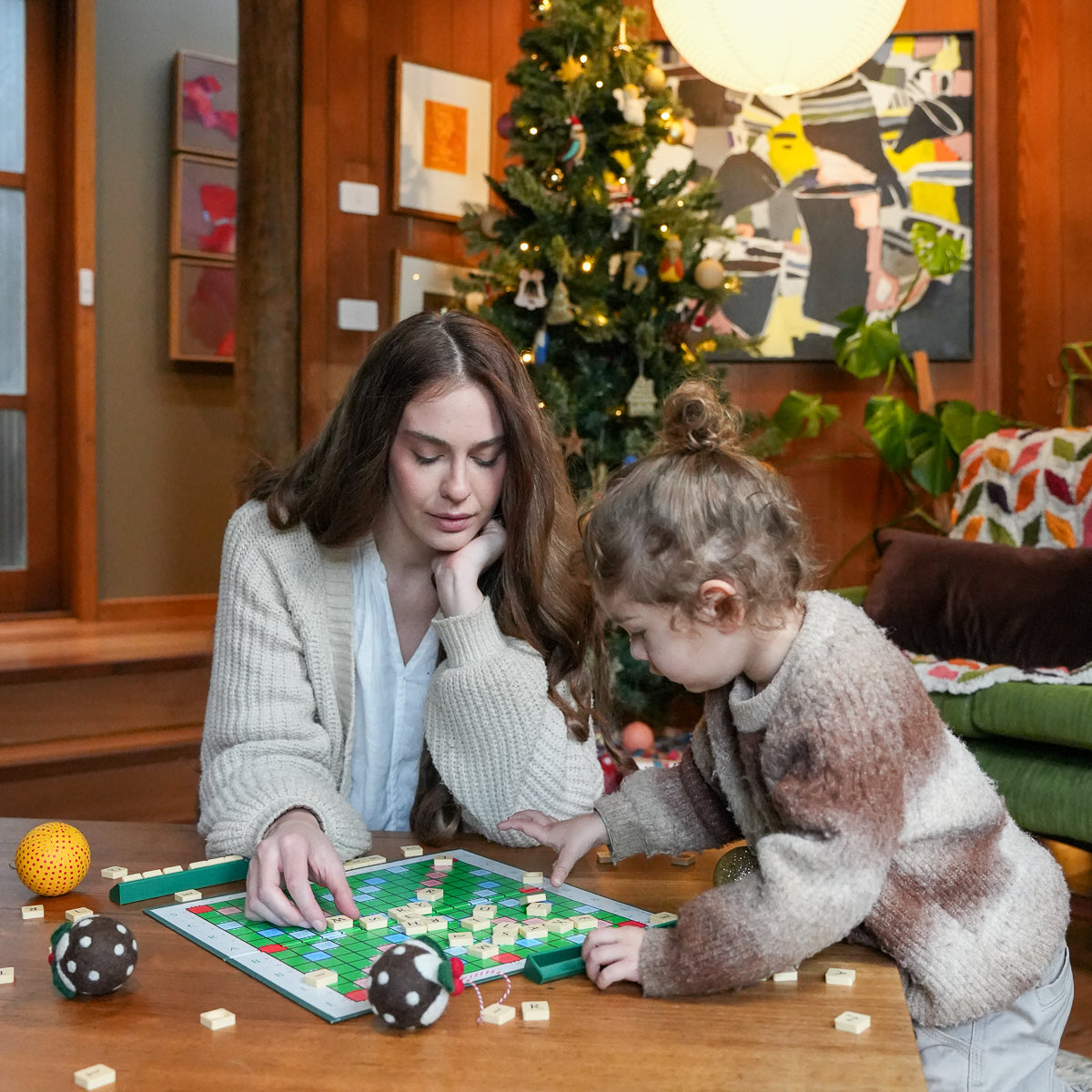 A woman and a young child are sitting at a wooden table playing with a board game in a cozy, decorated living room with a Christmas tree in the background.