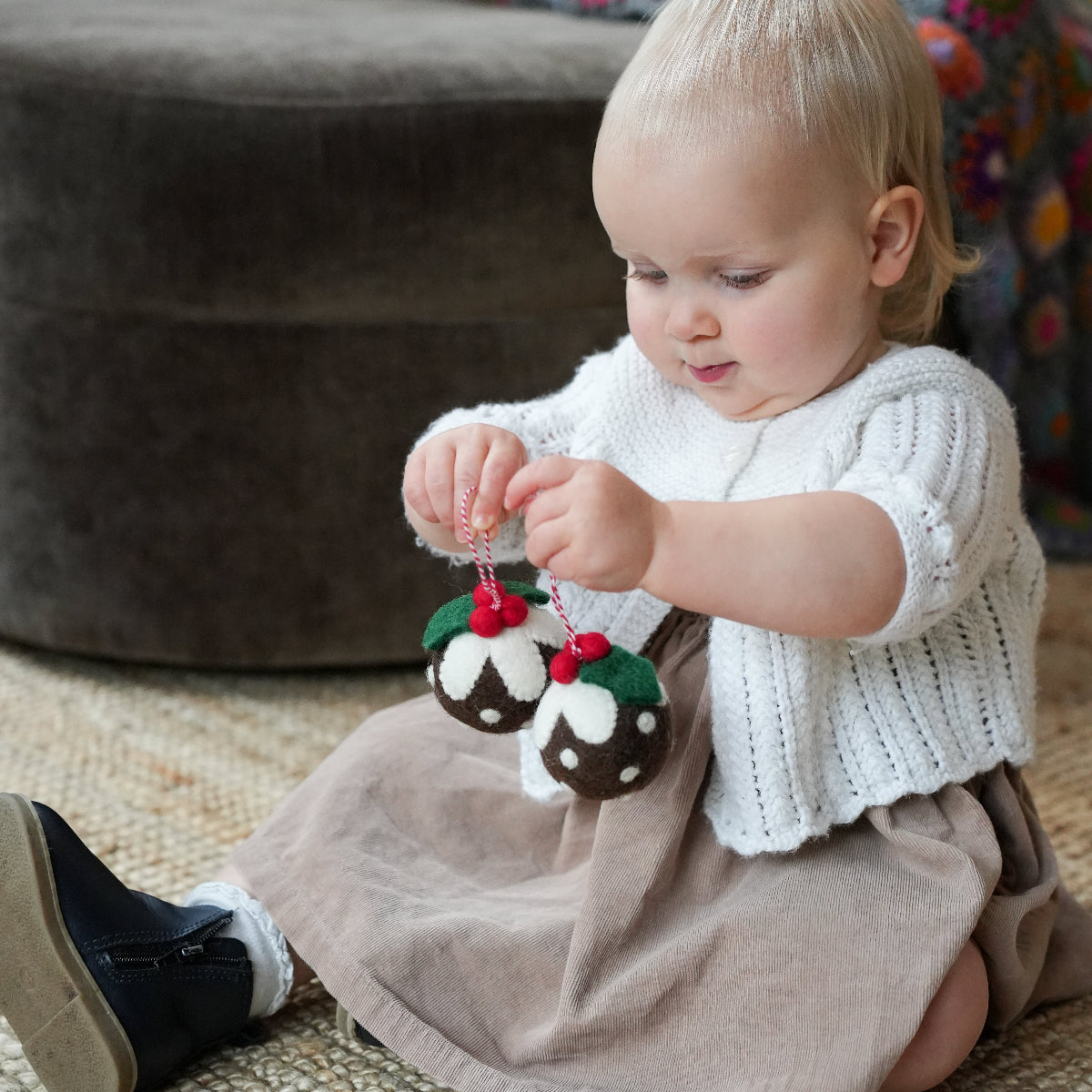 A toddler sits on a woven rug indoors, holding a small knitted ornament shaped like a Christmas nut with red berries. The child is wearing a white knit top and a taupe skirt, with a dark upholstered ottoman in the background.