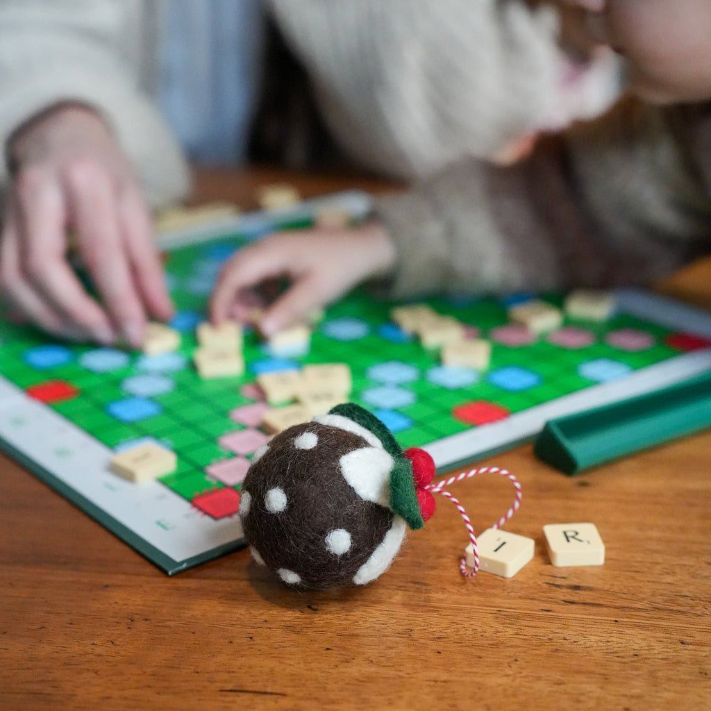 A close-up of a brown and white felt Christmas ornament in the foreground on a wooden table, with a person's hands and face blurred in the background playing with a green Scrabble board.