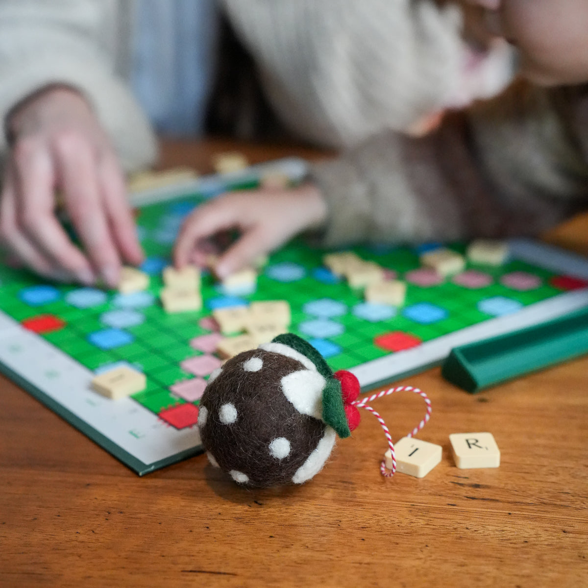 A close-up of a brown and white felt Christmas ornament in the foreground on a wooden table, with a person's hands and face blurred in the background playing with a green Scrabble board.