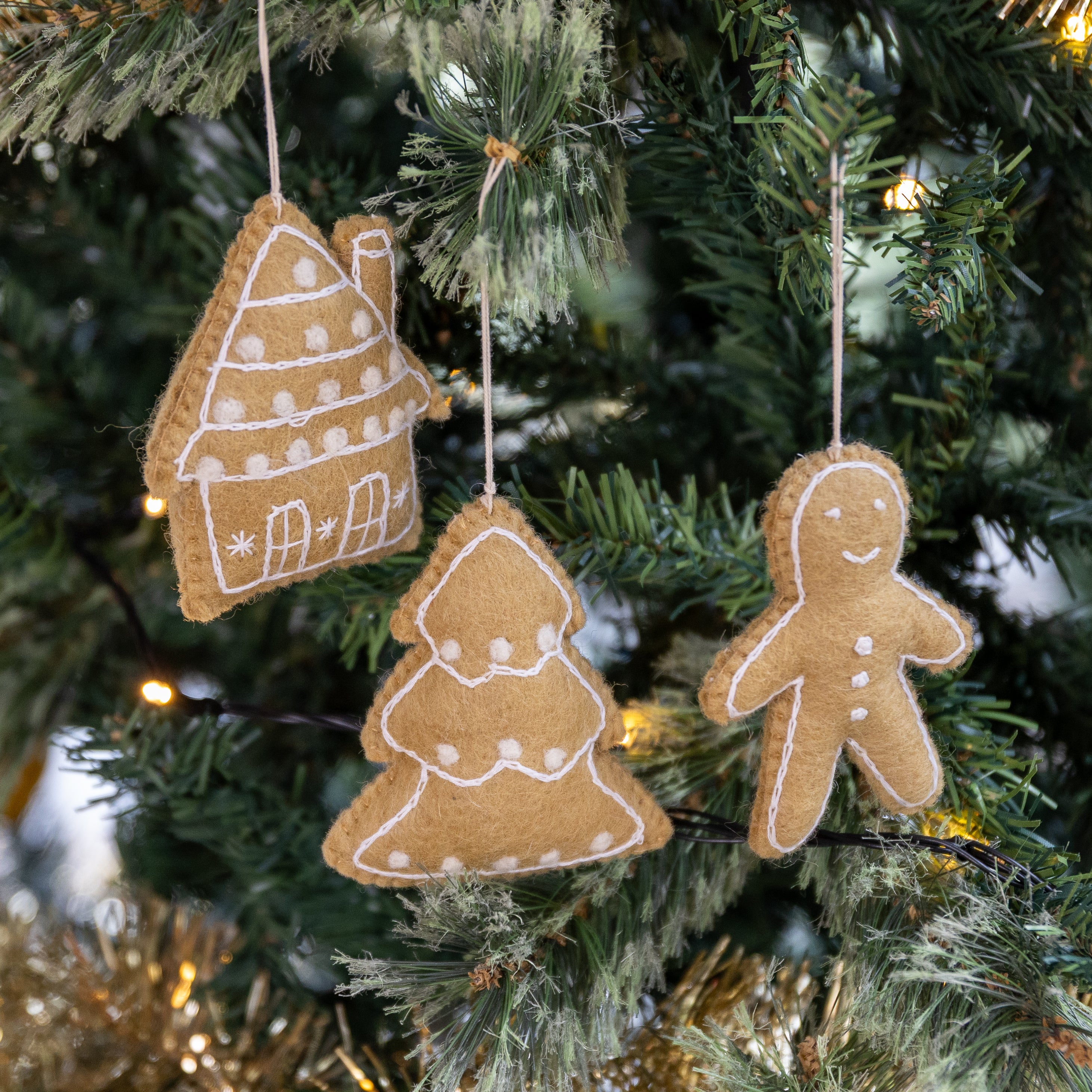 Three felt Christmas tree ornaments in the shapes of a house, a Christmas tree, and a gingerbread man, hanging on a decorated green Christmas tree with warm lights.