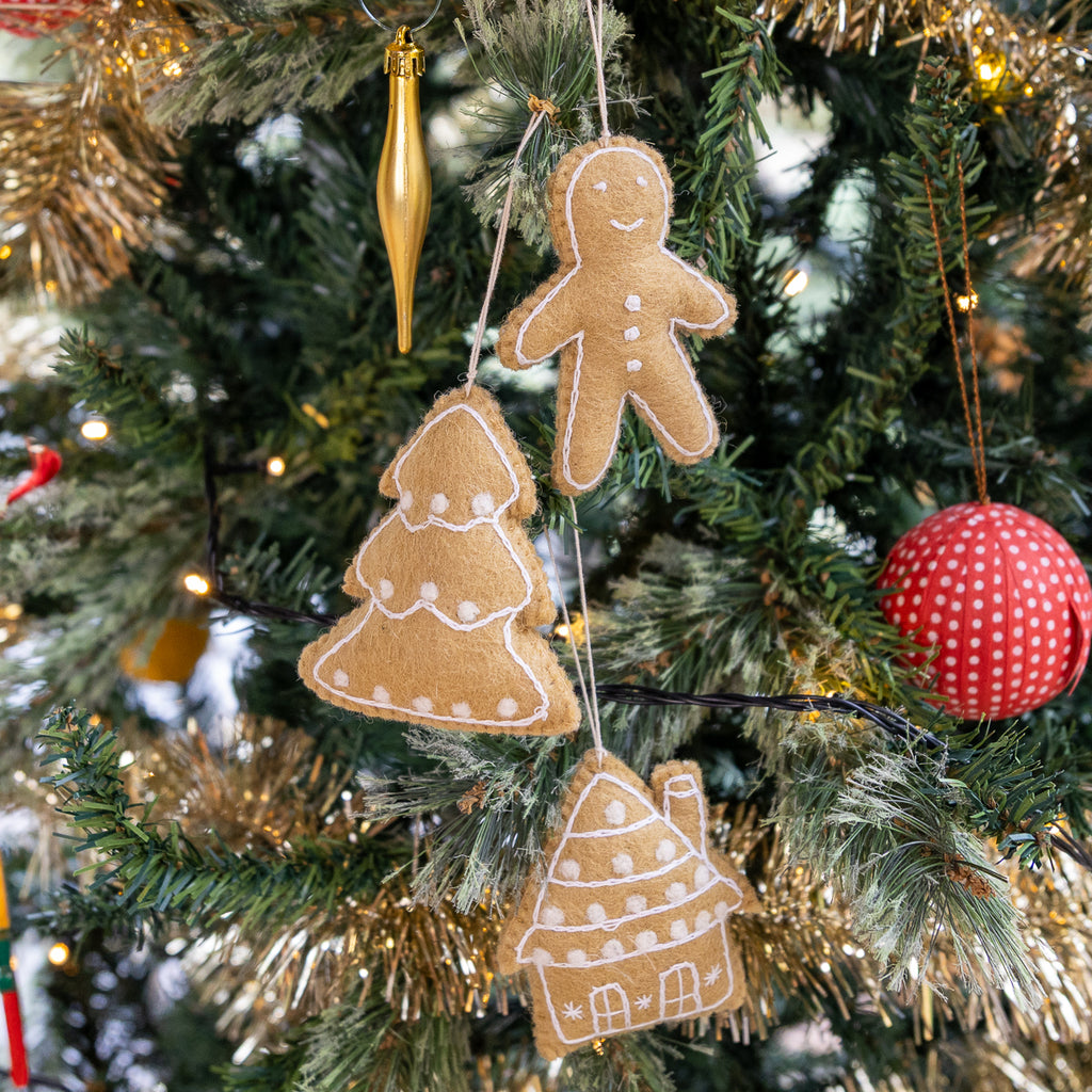 Three handmade felt Christmas ornaments shaped like a gingerbread man, tree, and house, decorated with white icing-like details and hanging on a decorated Christmas tree with lights.