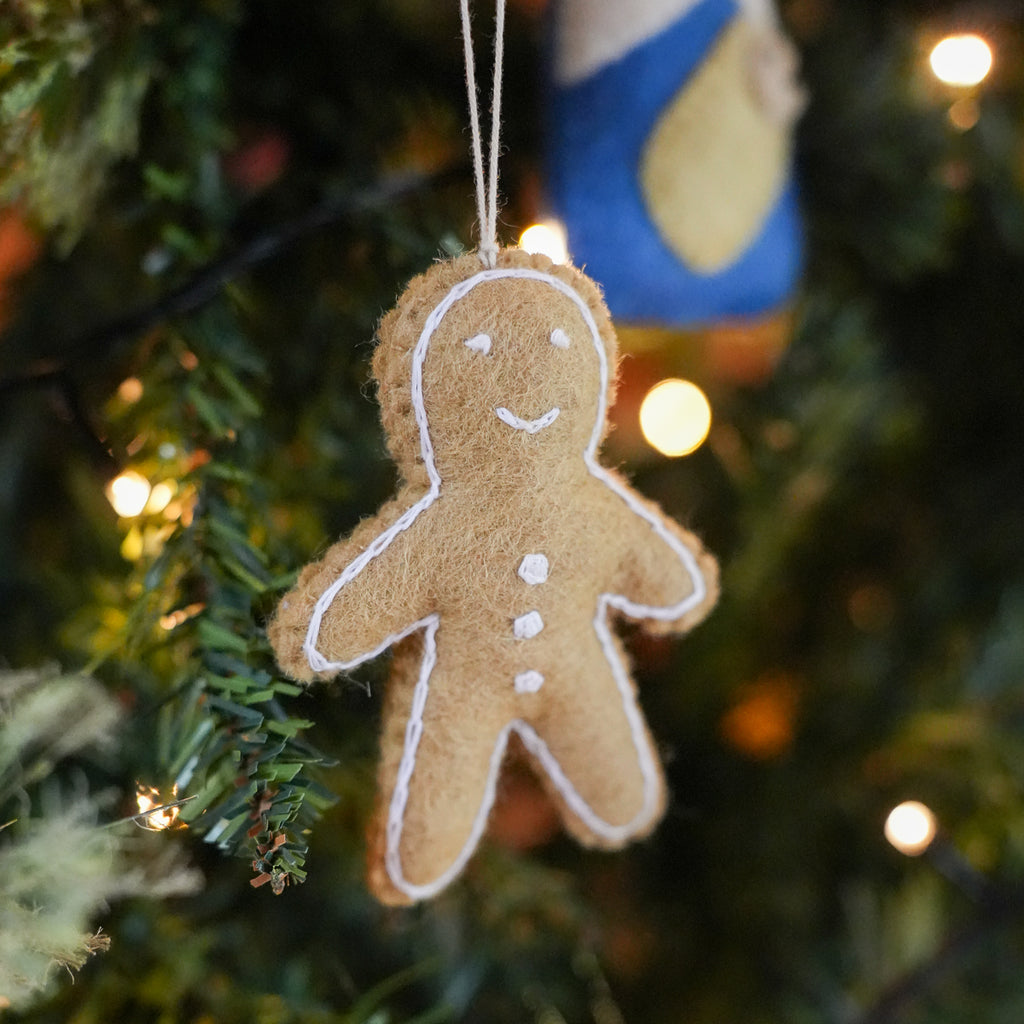A brown felt Christmas ornament shaped like a gingerbread man with white embroidered details, hanging on a decorated Christmas tree with warm lights.