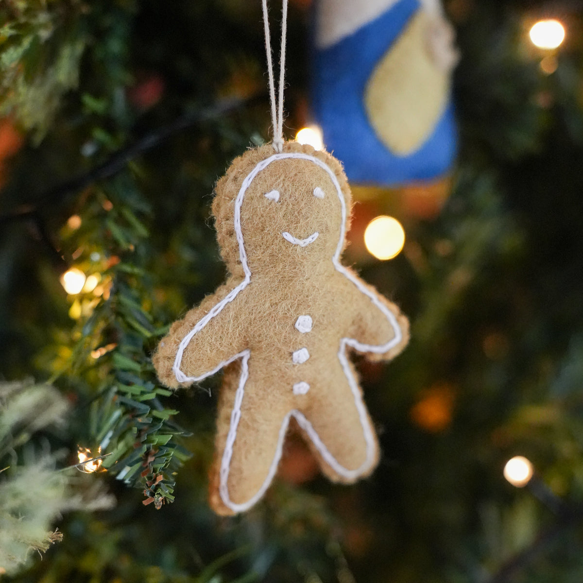 A brown felt Christmas ornament shaped like a gingerbread man with white embroidered details, hanging on a decorated Christmas tree with warm lights.