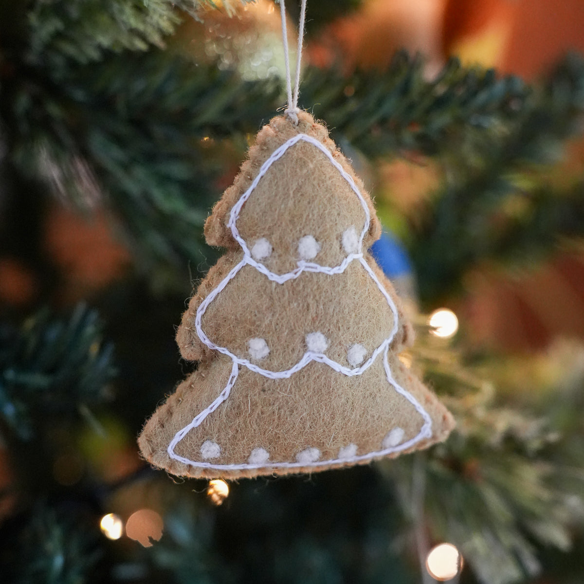 A felt Christmas tree ornament in a natural tan color with white stitched outlines and small white pom-pom details, hanging on a blurred evergreen Christmas tree with warm bokeh lights.