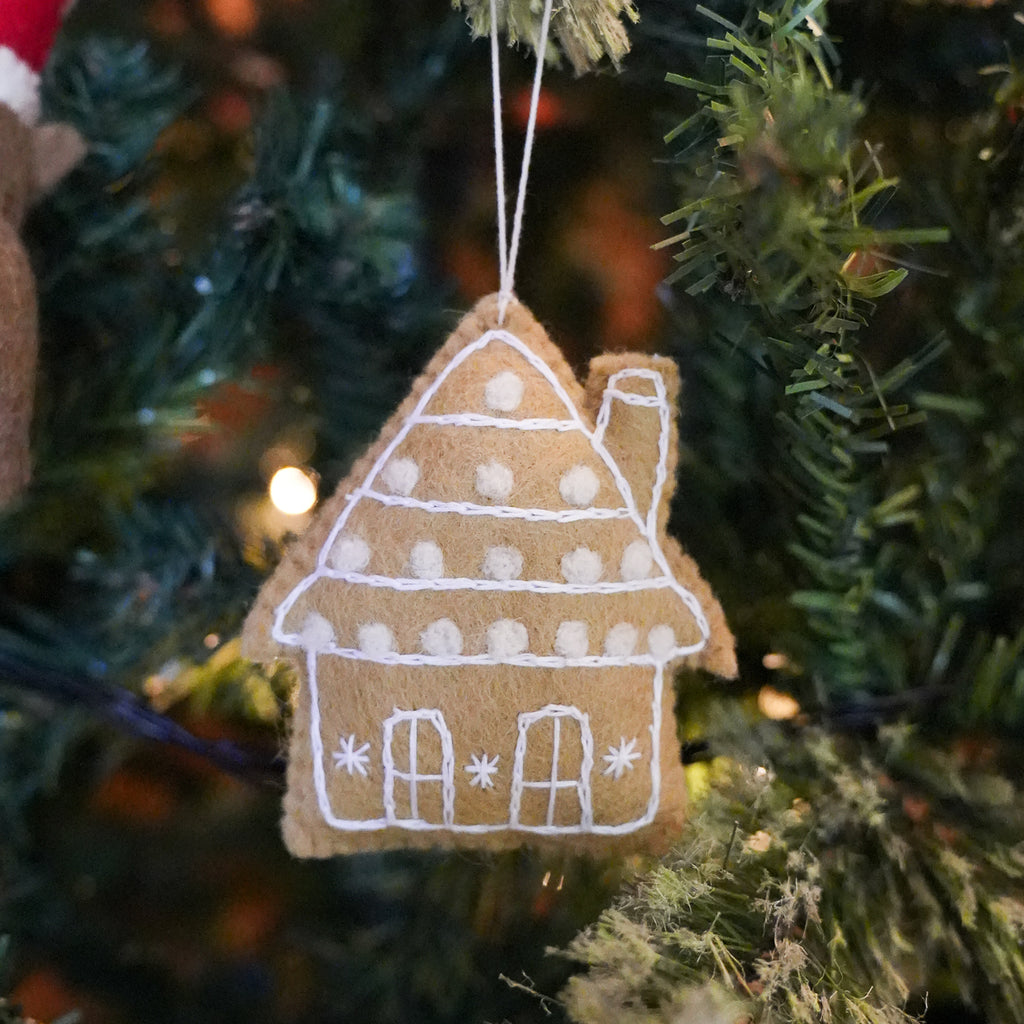 A felt Christmas house ornament in a warm tan color with white embroidered details, hanging from a green Christmas tree with warm lights in the background.