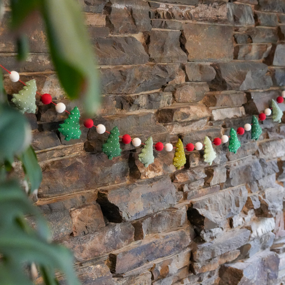 A festive Christmas garland featuring felt Christmas trees in green, light green, yellow, and white, interspersed with red and white pom-poms, hanging against a rustic stone wall with blurred green leaves in the foreground.