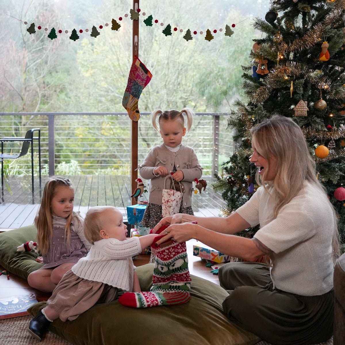 A woman and two young children are gathered on a floor cushion in a decorated holiday home, wrapping a red, green, and white Christmas stocking. A Christmas tree, window view, and festive garlands are visible in the background.
