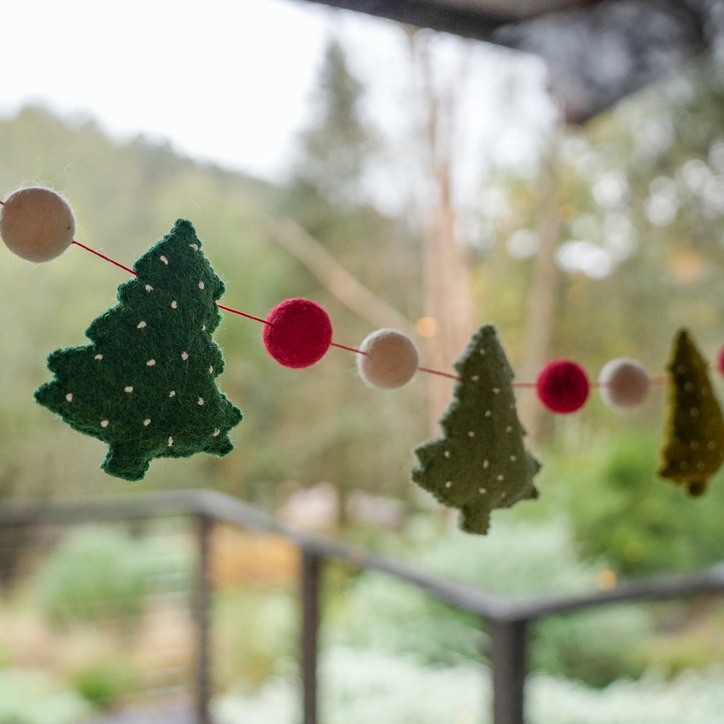 Felt Christmas tree garland with green and olive tree shapes and red and cream pom-pom beads strung on a red cord, photographed outdoors with a blurred garden background.