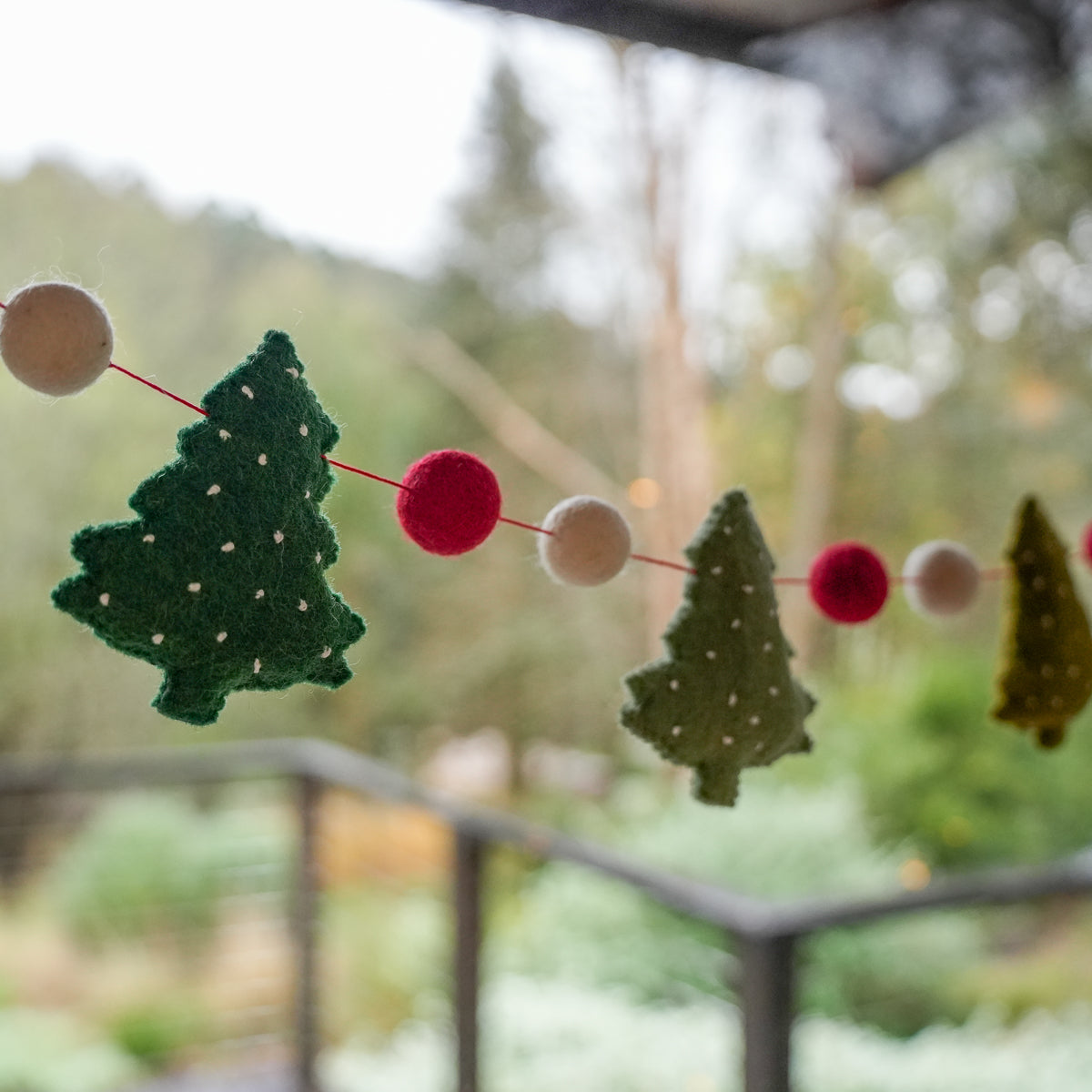 Felt Christmas tree garland with green and olive tree shapes and red and cream pom-pom beads strung on a red cord, photographed outdoors with a blurred garden background.