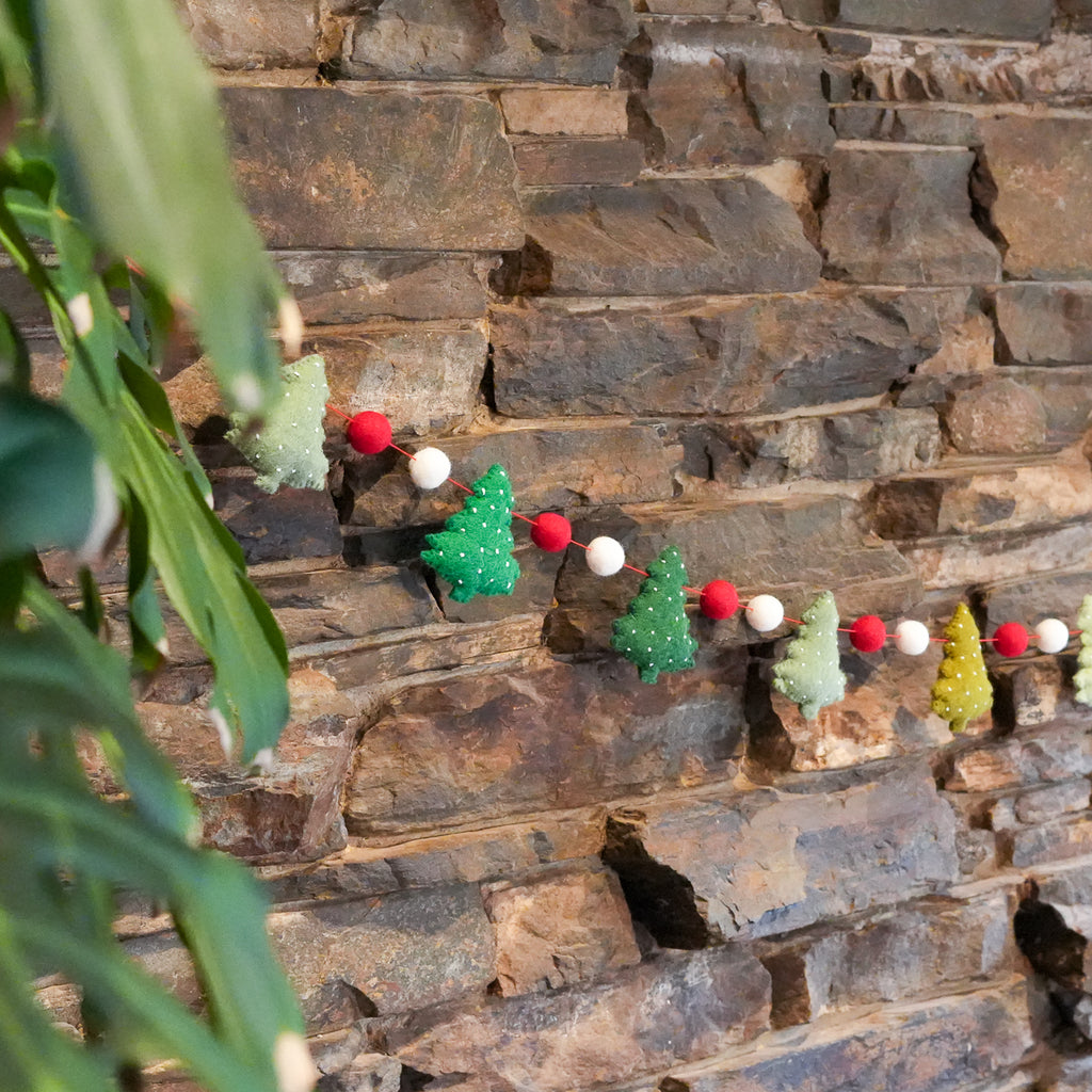 A festive felt garland with small Christmas trees and red-and-white pom-poms strung across a rustic stone wall, with green plant leaves in the foreground.