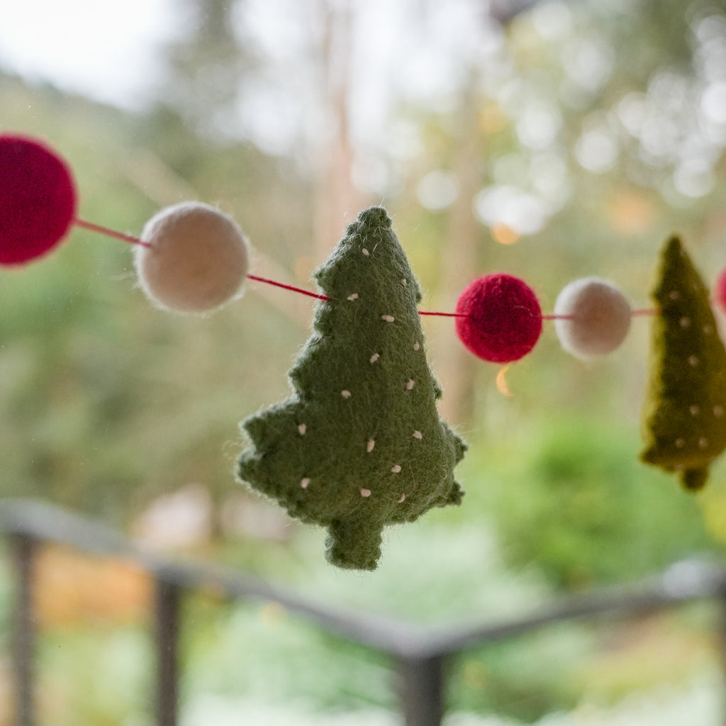 A felt holiday garland with green Christmas tree shapes and red and cream pom-poms strung across a window, with a blurred outdoor garden background.