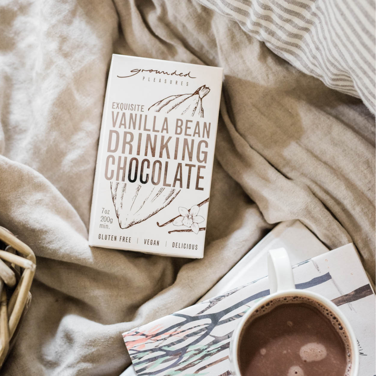 A box of Grounds Creek Vanilla Bean Drinking Chocolate is styled on a beige blanket next to a cup of hot chocolate and a book.