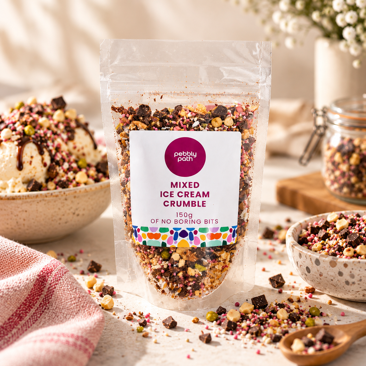 A clear stand-up pouch of Pebble Patch Mixed Ice Cream Crumble stands on a tabletop with bowls of the crumble and a spoon nearby, creating a styled food-product scene.