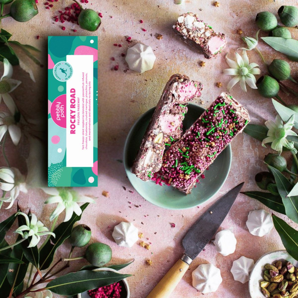 Two pink and white granola bars topped with pink sprinkles and green herbs sit on a light green plate, alongside their colorful packaging box labeled 'Planty Food Rocky Road'. The scene is styled as a flat lay with a wooden-handled knife, green olives, white meringues, and dried flowers on a textured pink surface.