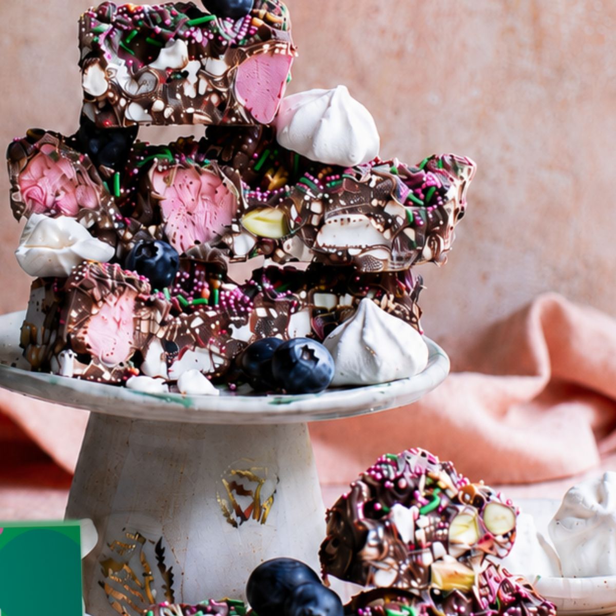 A close-up of assorted frozen dessert bars stacked on a white pedestal plate. The bars are topped with chocolate drizzle, sprinkles, blueberries, and white meringue-like dollops, with some bars broken in front on a pink fabric surface.