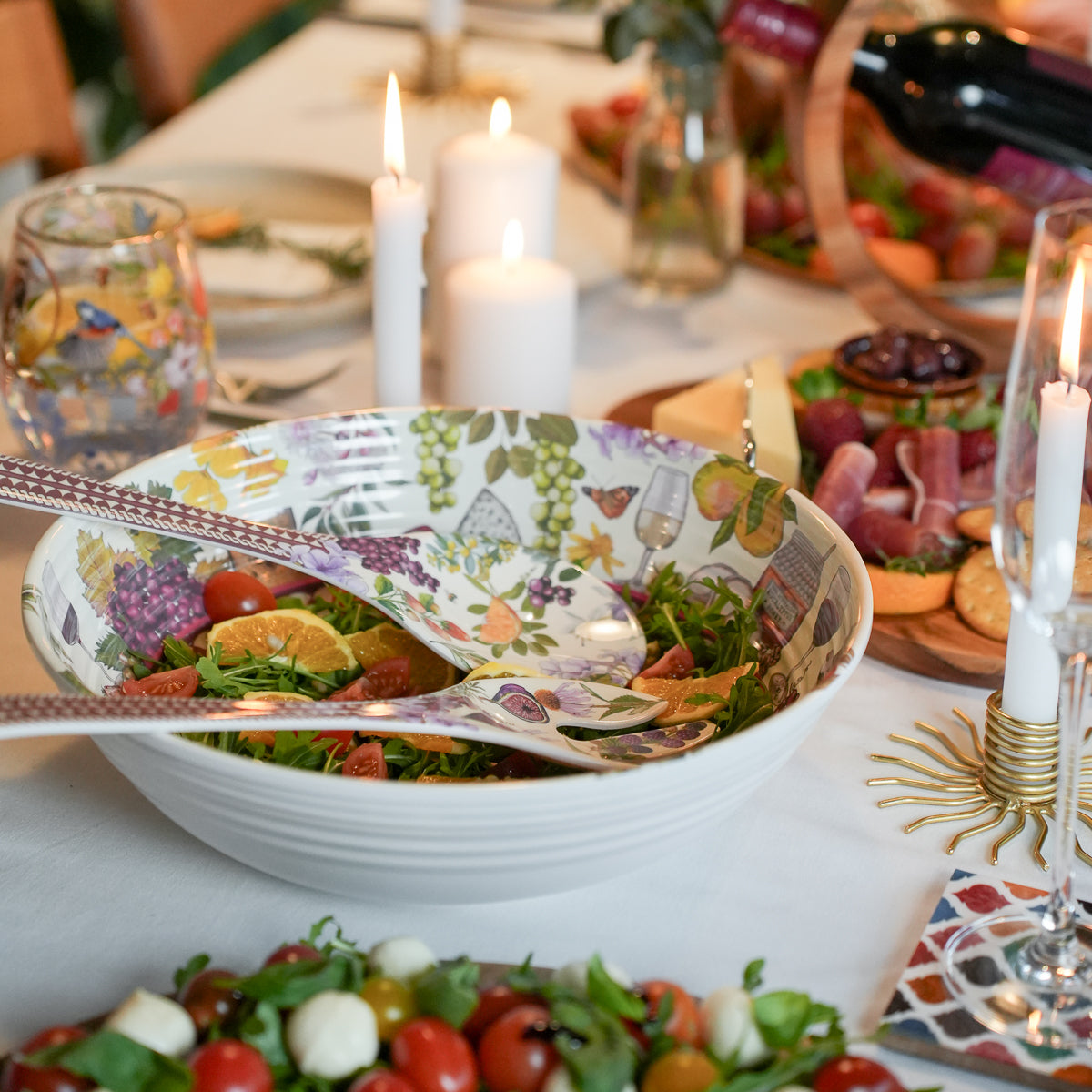 A large white ceramic salad bowl with a colorful floral and fruit pattern, filled with greens, tomatoes, and citrus slices, set on a decorated dining table with candles, charcuterie boards, and wine.