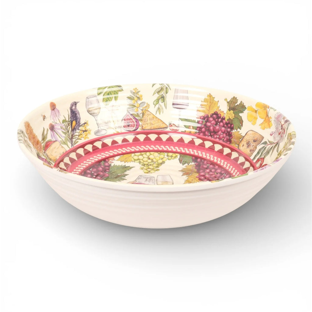 A round ceramic serving bowl with a white ribbed exterior and a detailed wine-themed interior pattern featuring red grapes, green leaves, wine glasses, and a red geometric band.