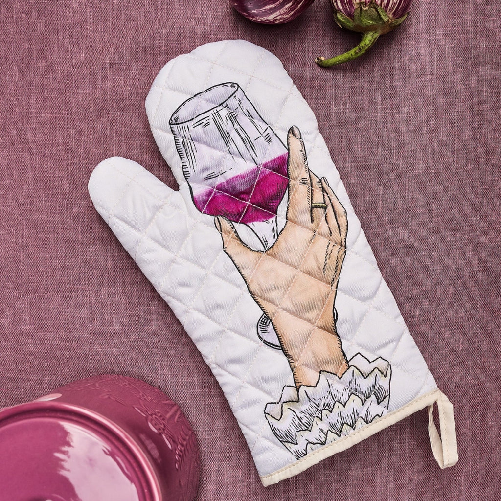 A white quilted oven mitt featuring an illustration of a hand holding a glass of red wine, laid flat on a textured mauve fabric background with kitchen props.