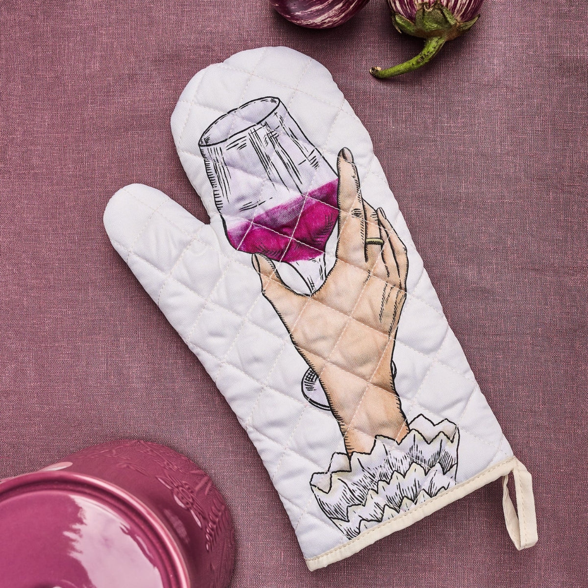 A white quilted oven mitt featuring an illustration of a hand holding a glass of red wine, laid flat on a textured mauve fabric background with kitchen props.