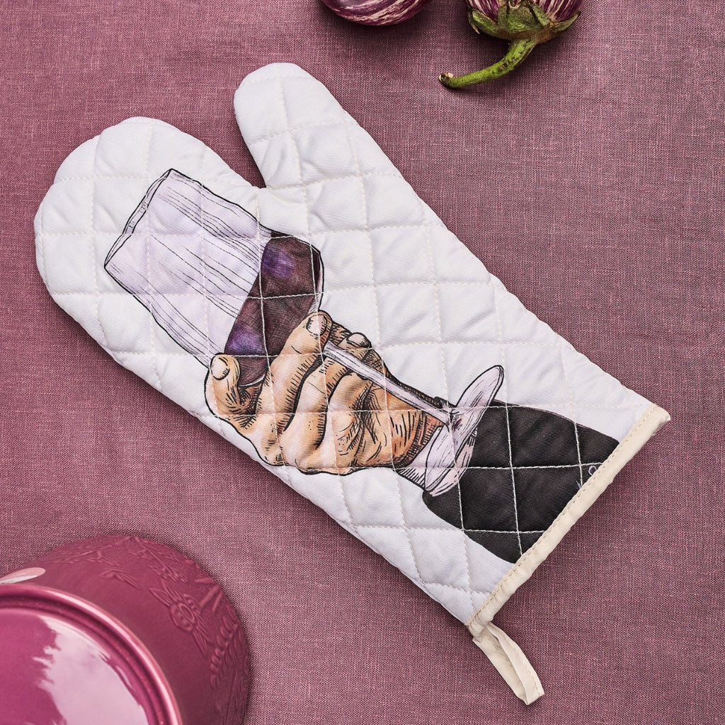 A quilted white oven mitt with a printed illustration of a hand holding a wine glass, placed on a textured mauve fabric background with kitchen props partially visible.