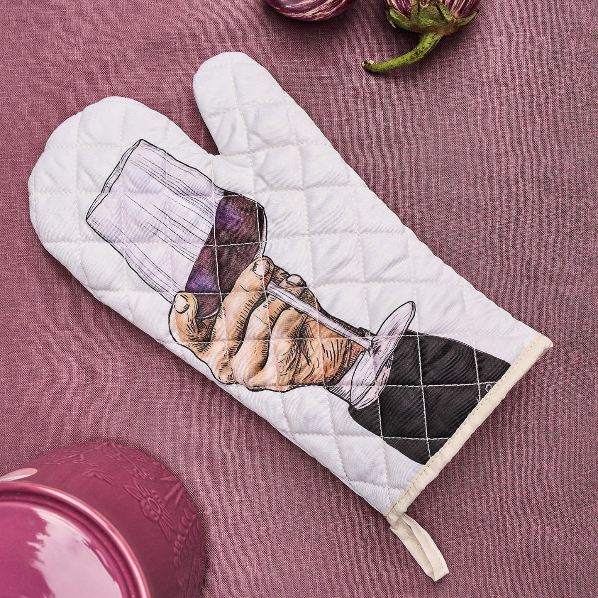 A quilted white oven mitt with a printed illustration of a hand holding a wine glass, placed on a textured mauve fabric background with kitchen props partially visible.